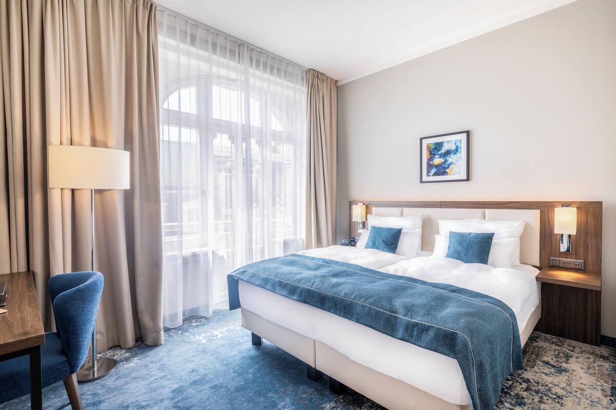 Une chambre d’hôtel avec un grand lit, un bureau et une chaise. Il y a une fenêtre avec des rideaux, un tableau au mur et une lampe sur la table de chevet.