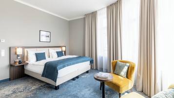 Ein Hotelzimmer mit einem Bett, einem kleinen runden Tisch, einem gelben Sessel und einem großen Fenster mit Vorhängen.