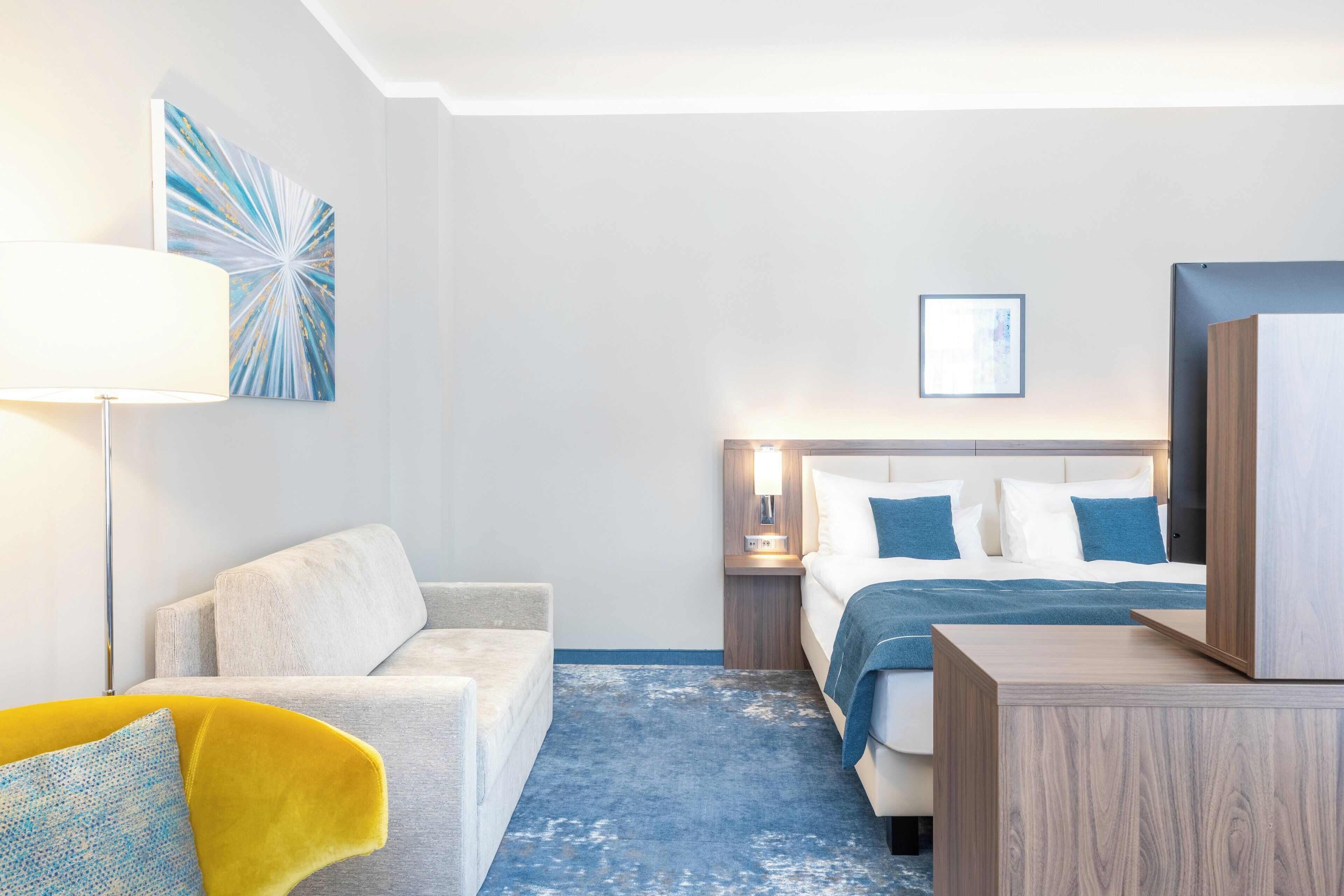 A modern hotel room with a bed, a sofa, a desk, and a lamp.