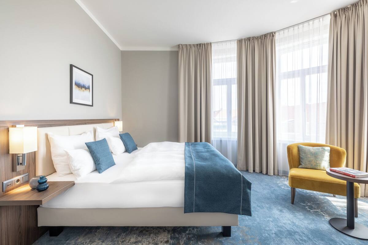 Une chambre d’hôtel avec un grand lit, une table de chevet avec une lampe, une chaise et une fenêtre avec des rideaux.