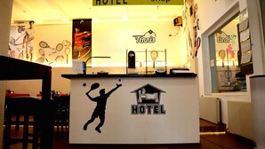 Een receptiebalie met een silhouet van een tennisser, een bord met de naam Tennis Hotel, en een ontvangstgedeelte met een balie en stoelen.