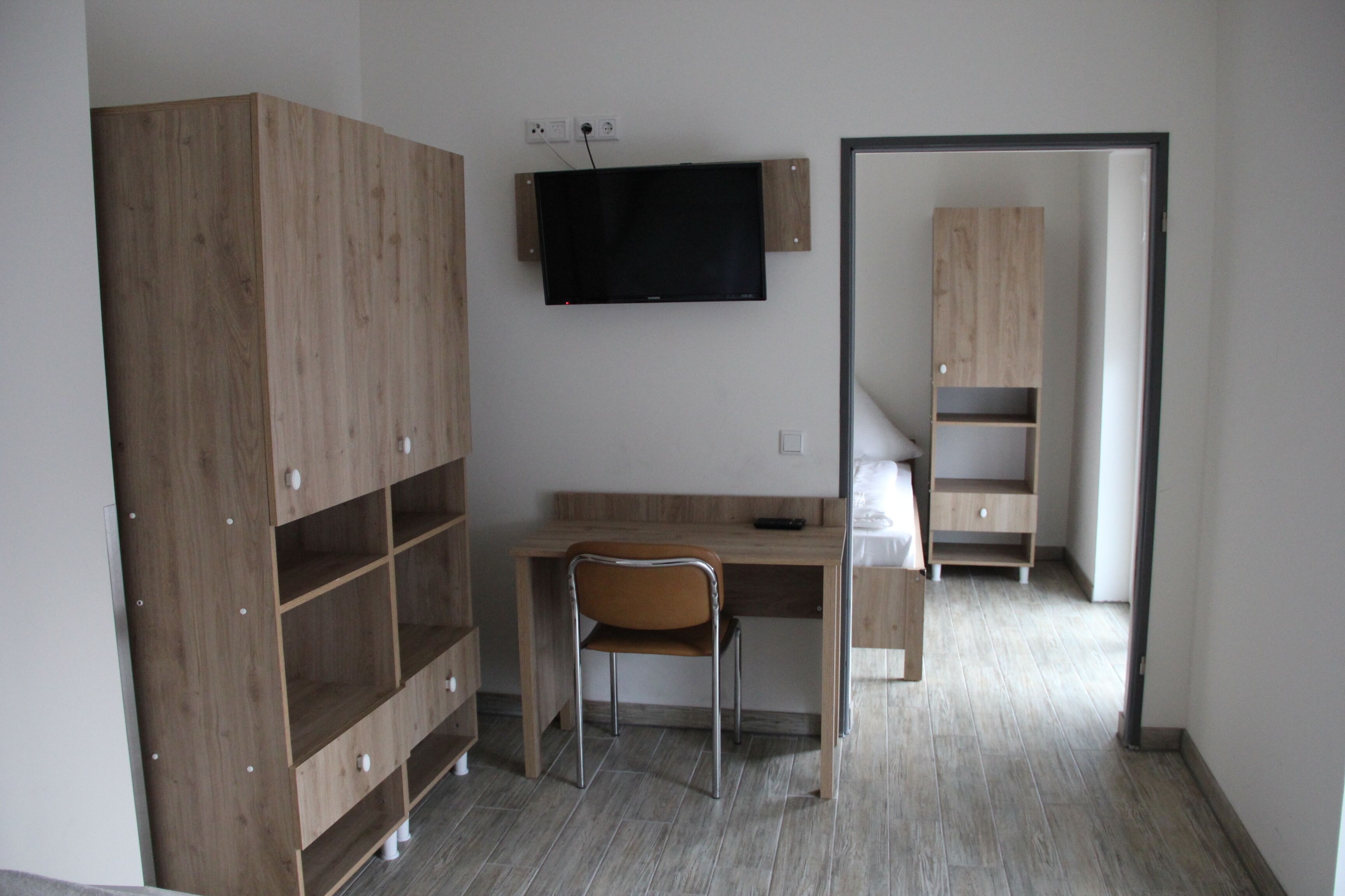 A room with a wooden desk, a chair, a television mounted on the wall, and a wardrobe with multiple shelves.