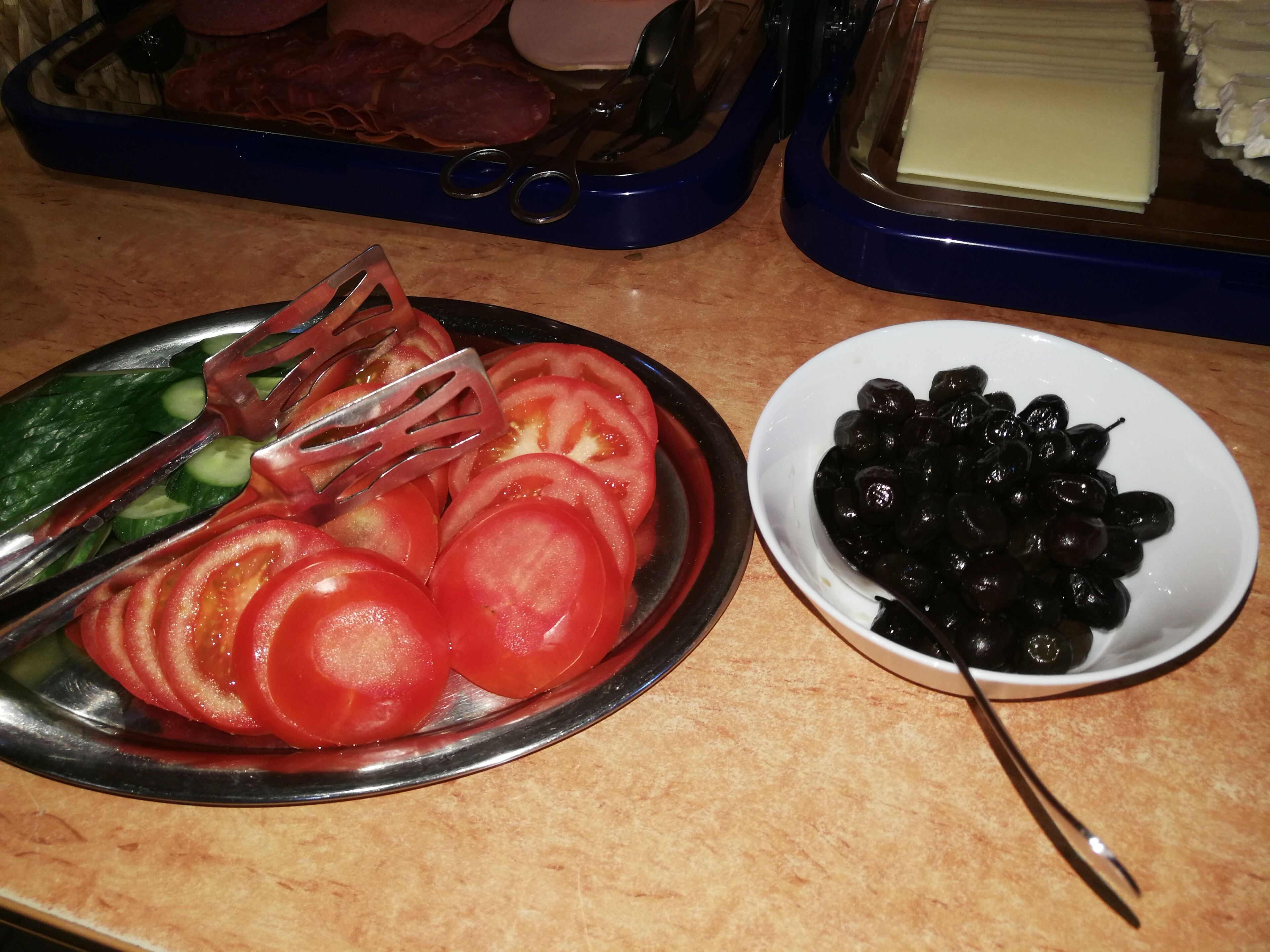 Ein Teller mit Tomaten- und Gurkenscheiben, eine Gabel, eine Schüssel schwarze Oliven und eine Platte mit Aufschnitt.