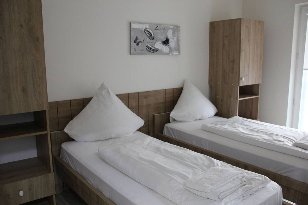 A room with two single beds, wooden headboards, white bedding, a wooden wardrobe, and a wall-mounted artwork.