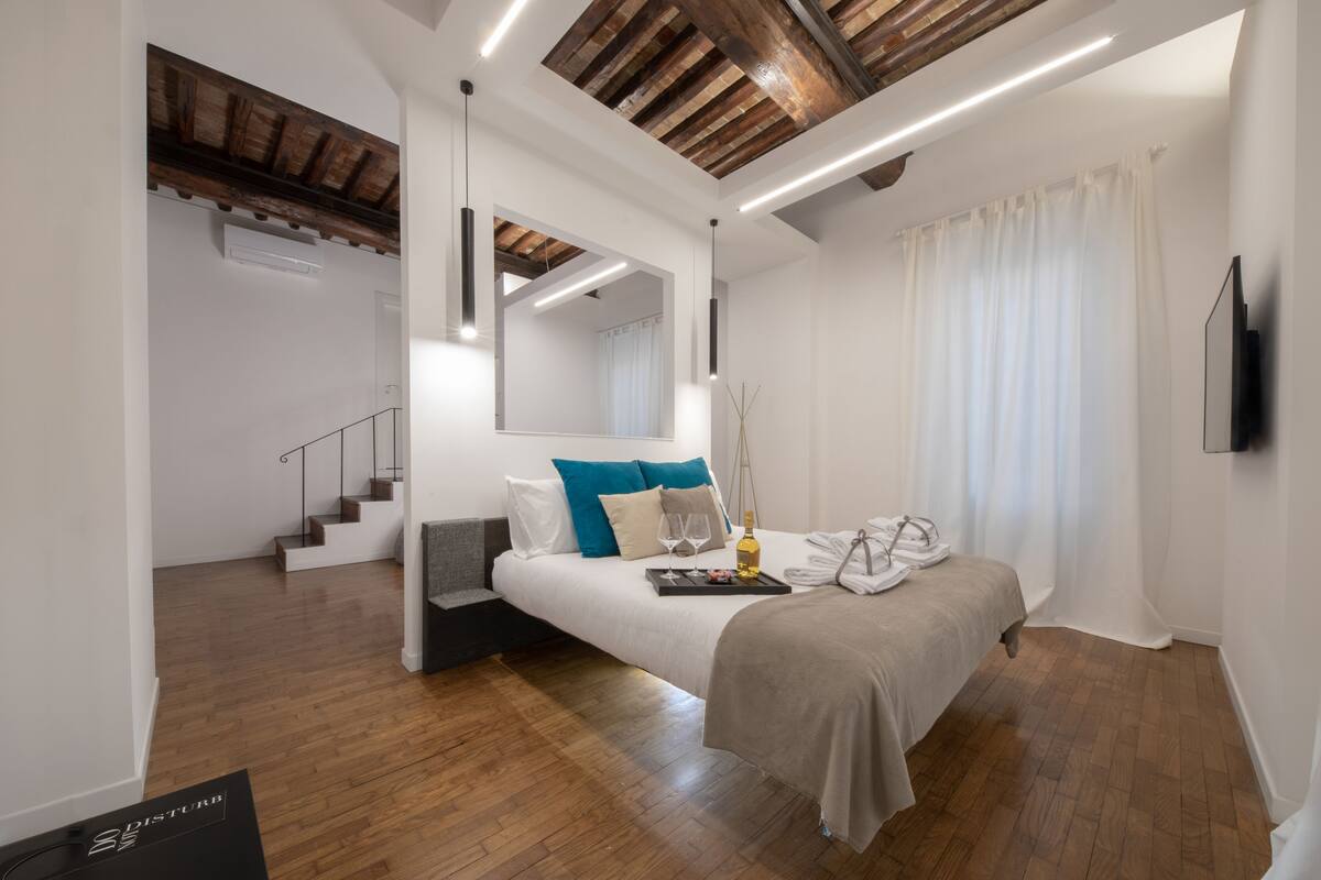 A modern bedroom with a wooden ceiling, a large bed with a tray of glasses, a TV, and a staircase leading to another room.