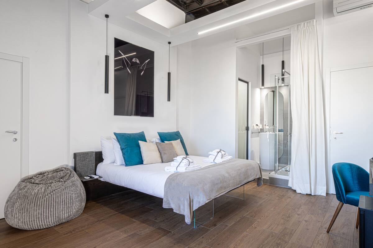 A modern bedroom with a bed, a bedside table, a chair, a large mirror, and a shower area.