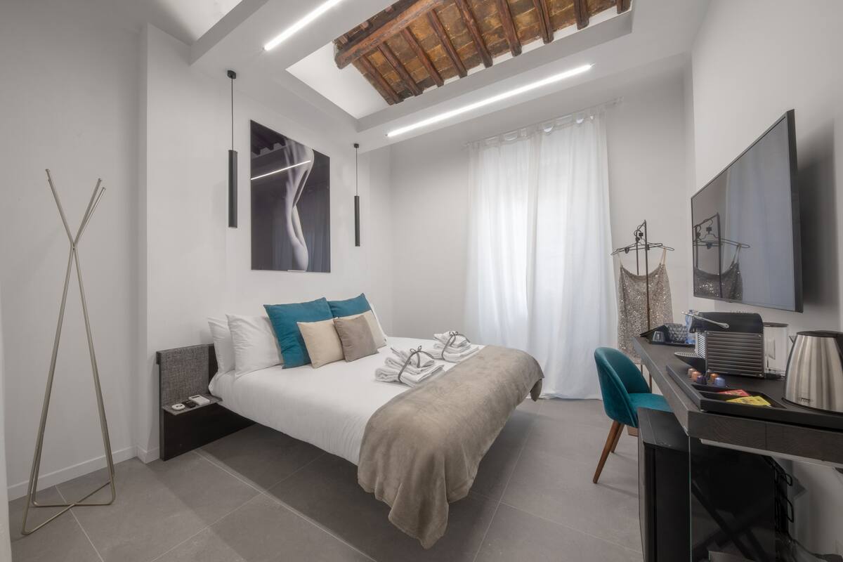 A modern hotel room with a bed, a desk, a chair, a TV, and a ceiling light fixture.