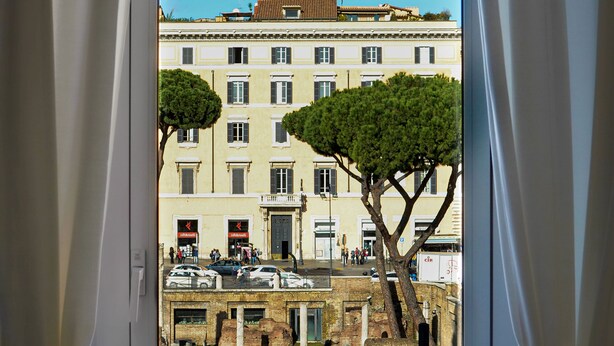 A window view of a building with a clear sky, trees, and a street with parked cars. - Argentina Suites (Rome)