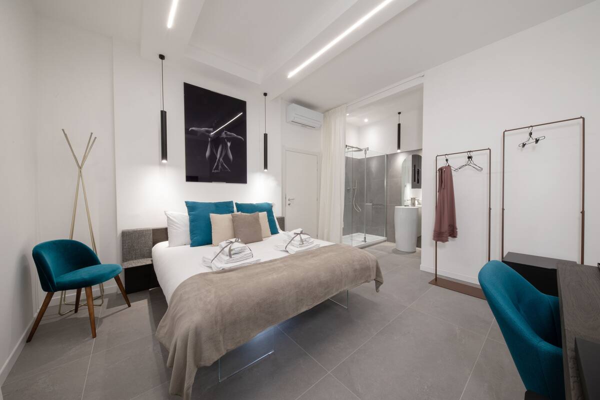 A modern hotel room with a large bed, a seating area with a chair, a mirror, and a shower area.