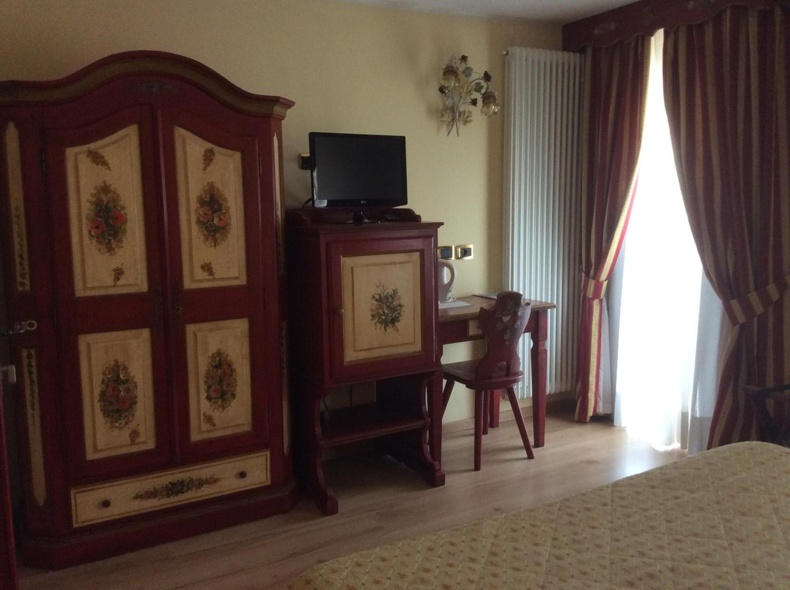 Una stanza con un letto, un'armadietto in legno decorato con motivi floreali, una scrivania con una sedia e un termosifone.