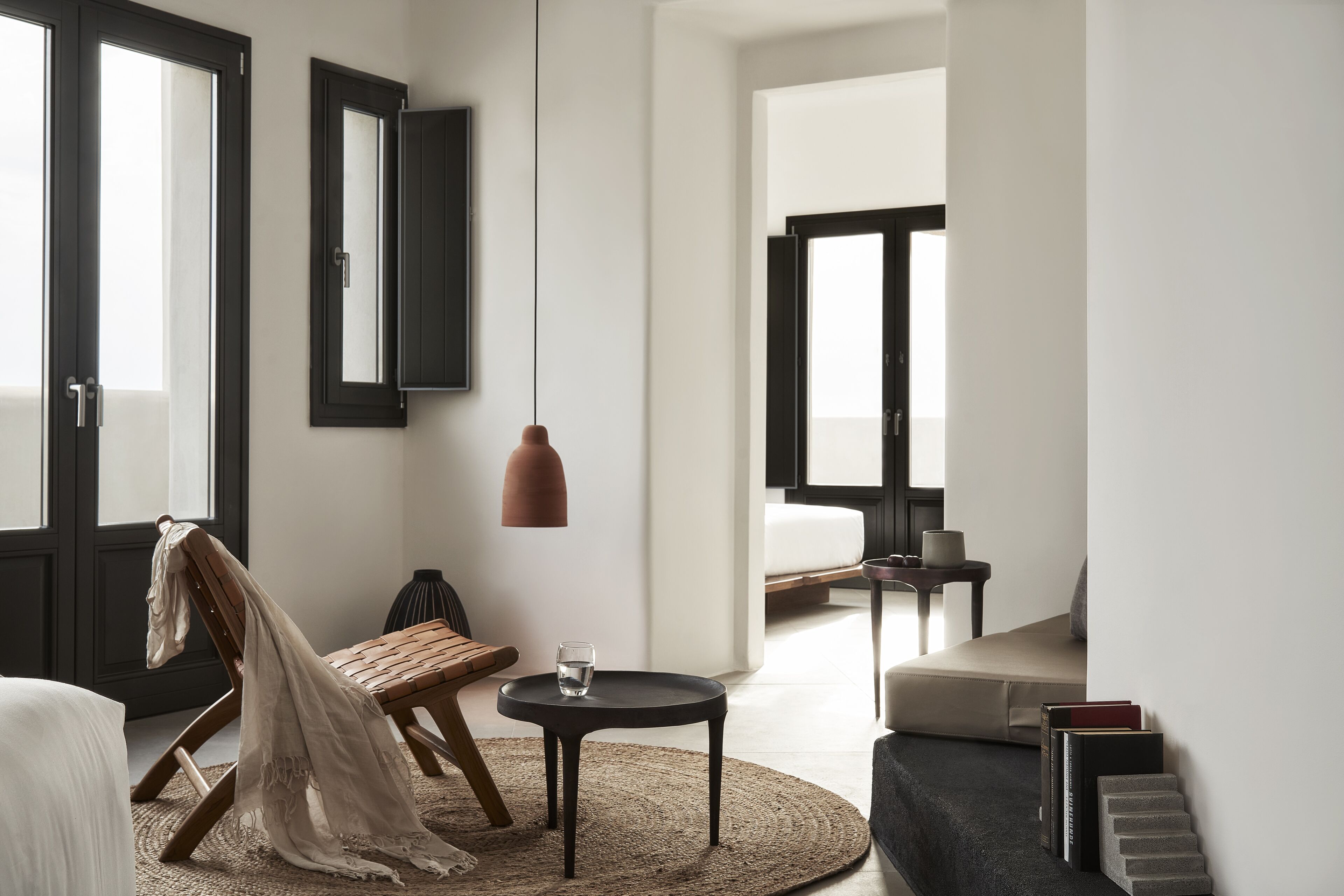 Una habitación de hotel moderna con una cama grande, una silla de madera, una mesita redonda con un vaso de agua, una lámpara colgante y vistas por una puerta abierta.