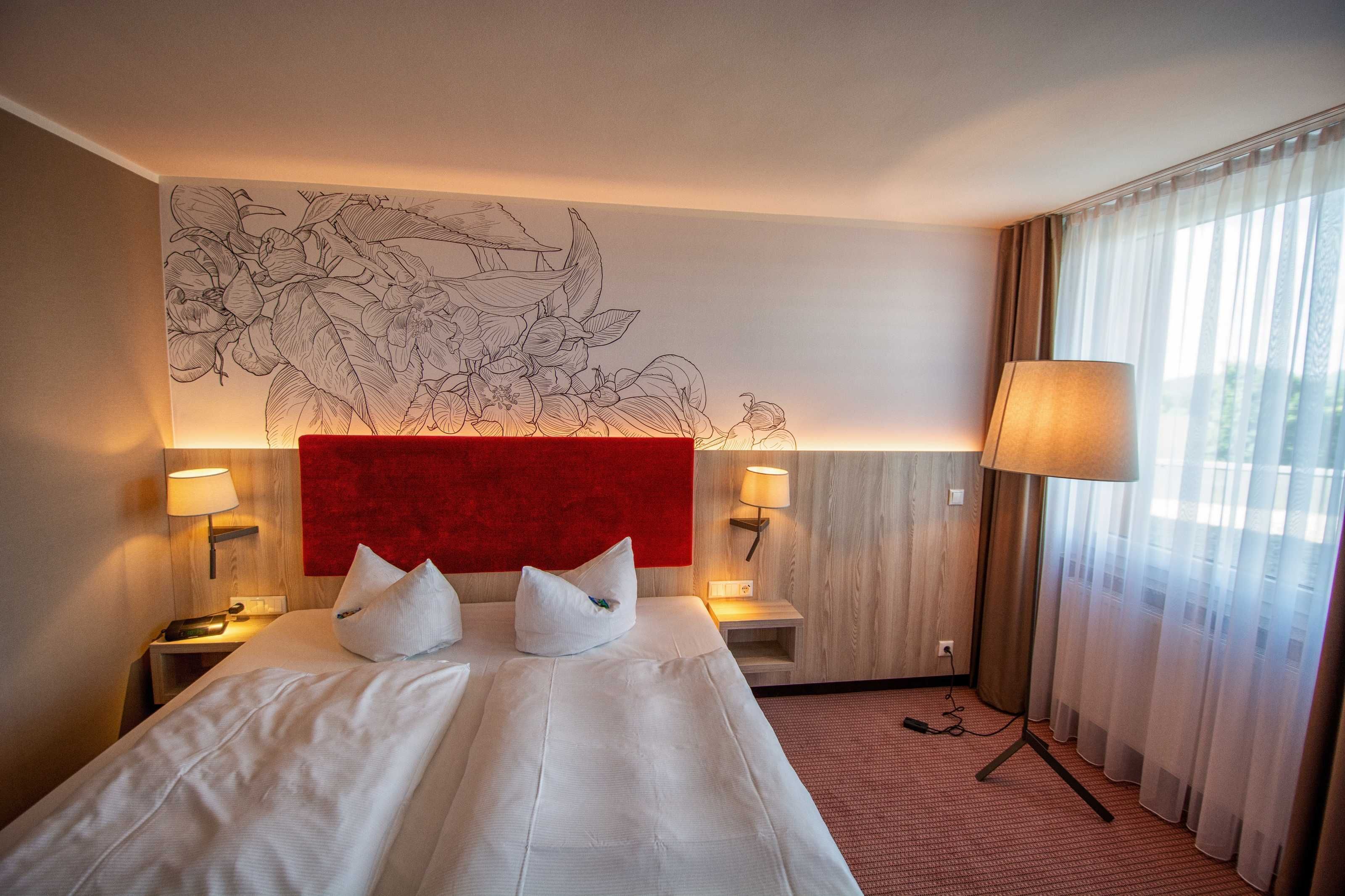A hotel room with a large bed, two bedside tables with lamps, a red accent wall, and a large window with sheer curtains.