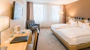 A hotel room with a bed, a desk with a laptop, a chair, a TV, and a window with curtains.