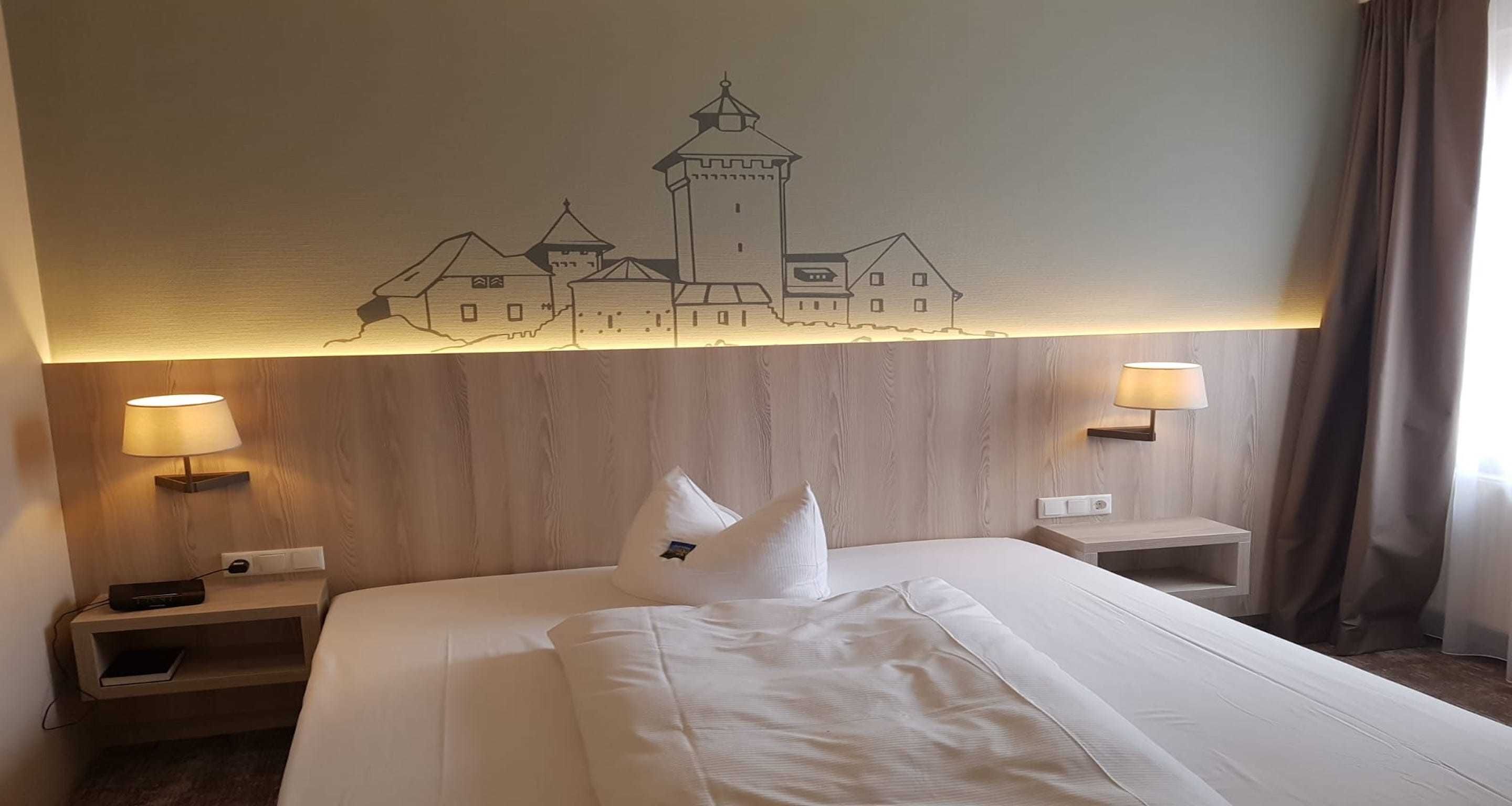 A hotel room with a large bed, two bedside tables, a wall-mounted lamp, and a wall art depicting a castle.