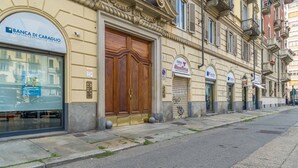Vista de calle con un edificio que tiene una puerta de madera grande, un escaparate con un letrero que dice 'Banca di Caraglio' y otro letrero que indica 'Cesa Macelleria'.