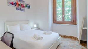 Habitación de hotel con cama, una silla, mesita de noche, lámpara, ventana y cuadros en la pared.