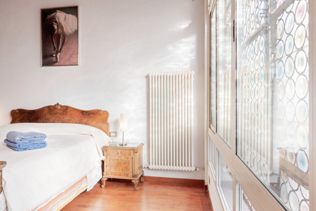 A bedroom with a wooden headboard bed, a nightstand with a lamp, a radiator, and a large window with a patterned view.