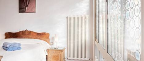 A bedroom with a wooden headboard bed, a nightstand with a lamp, a radiator, and a large window with a patterned view.