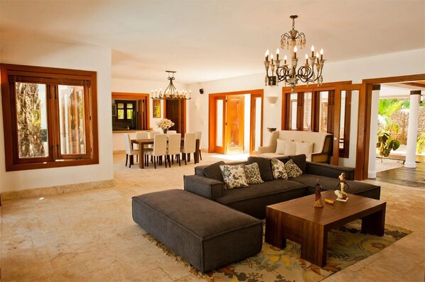 A spacious living room with a large sectional sofa, a coffee table, and a chandelier. There is a dining area with a table and chairs visible in the background.