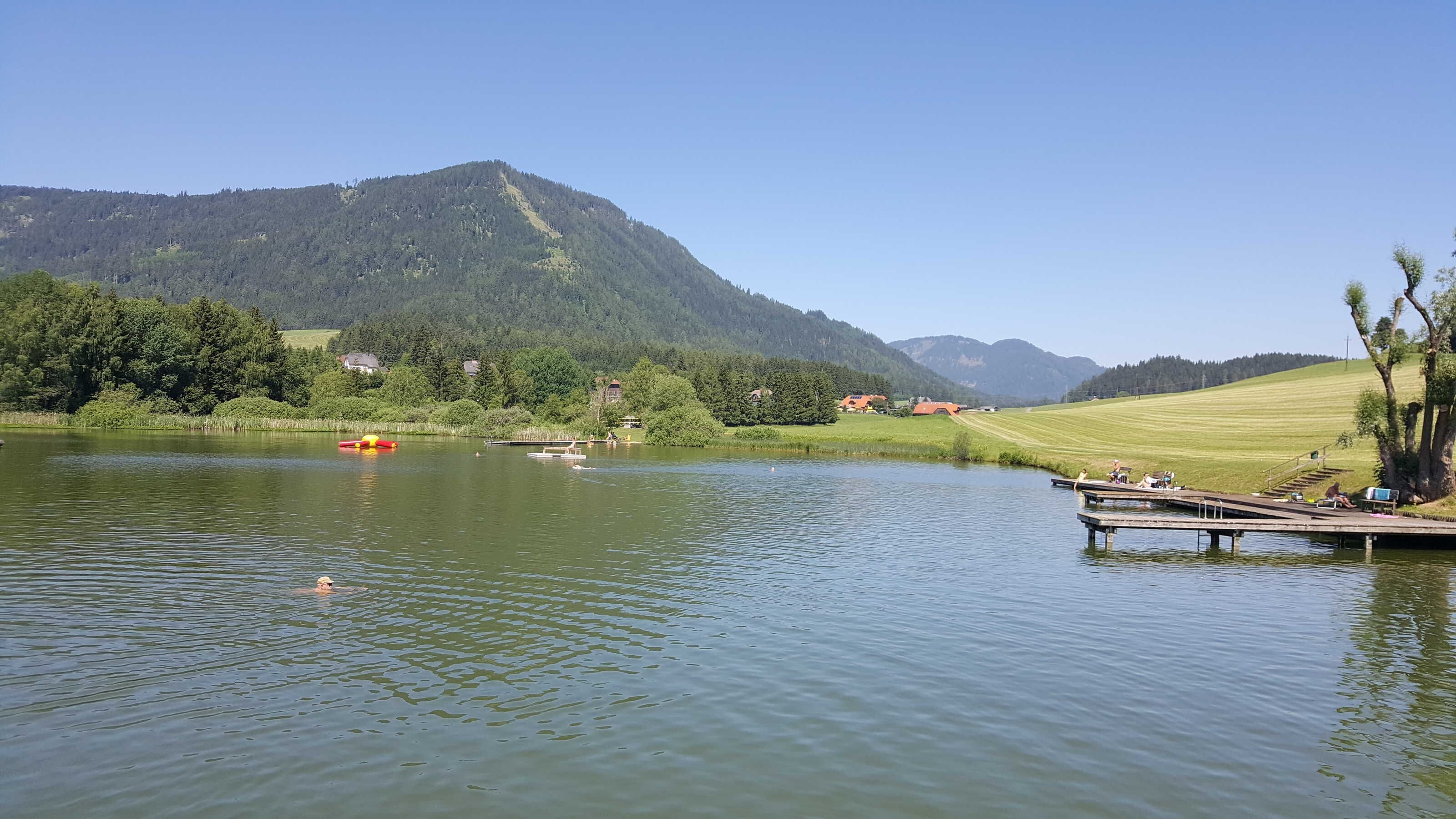 Un lac avec un quai, entouré de collines boisées et de maisons.