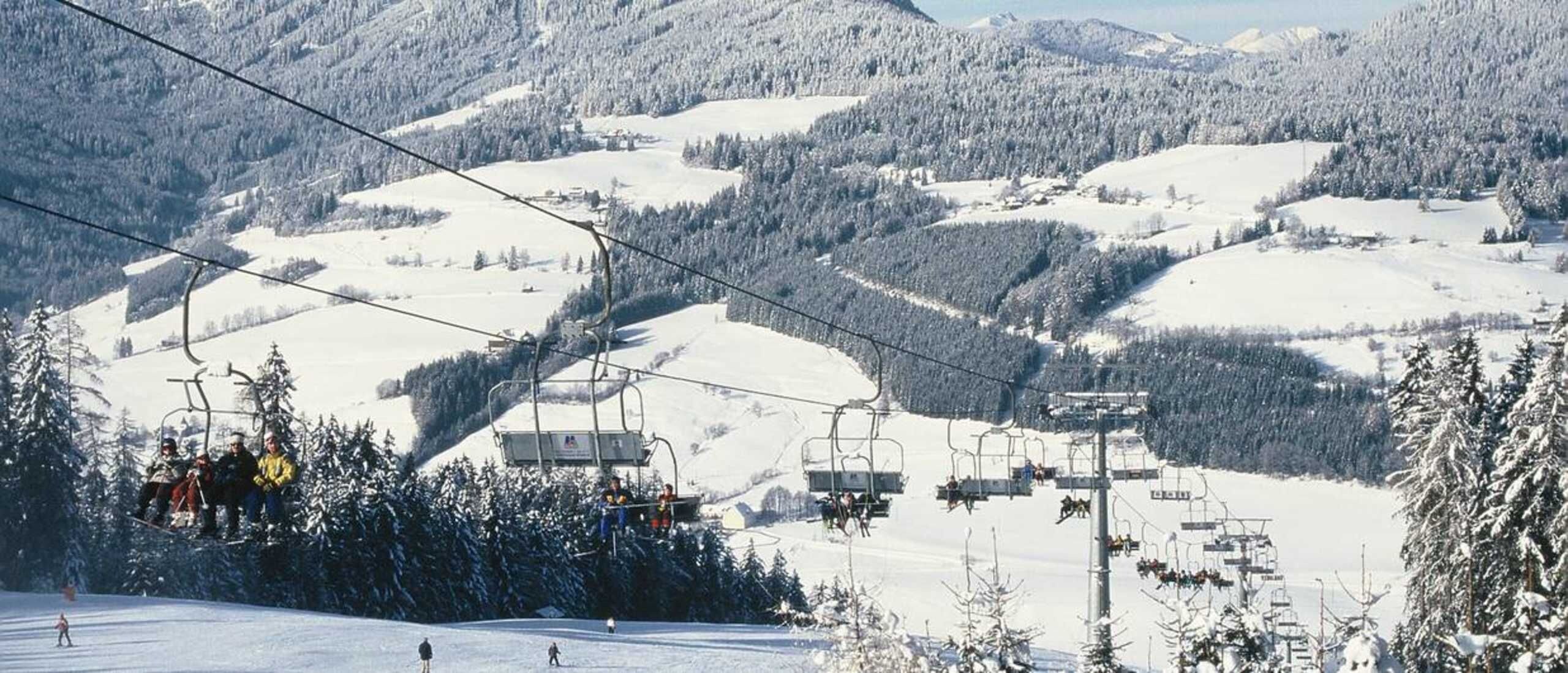 Un télésiège avec des skieurs, un paysage montagneux enneigé et une zone boisée.