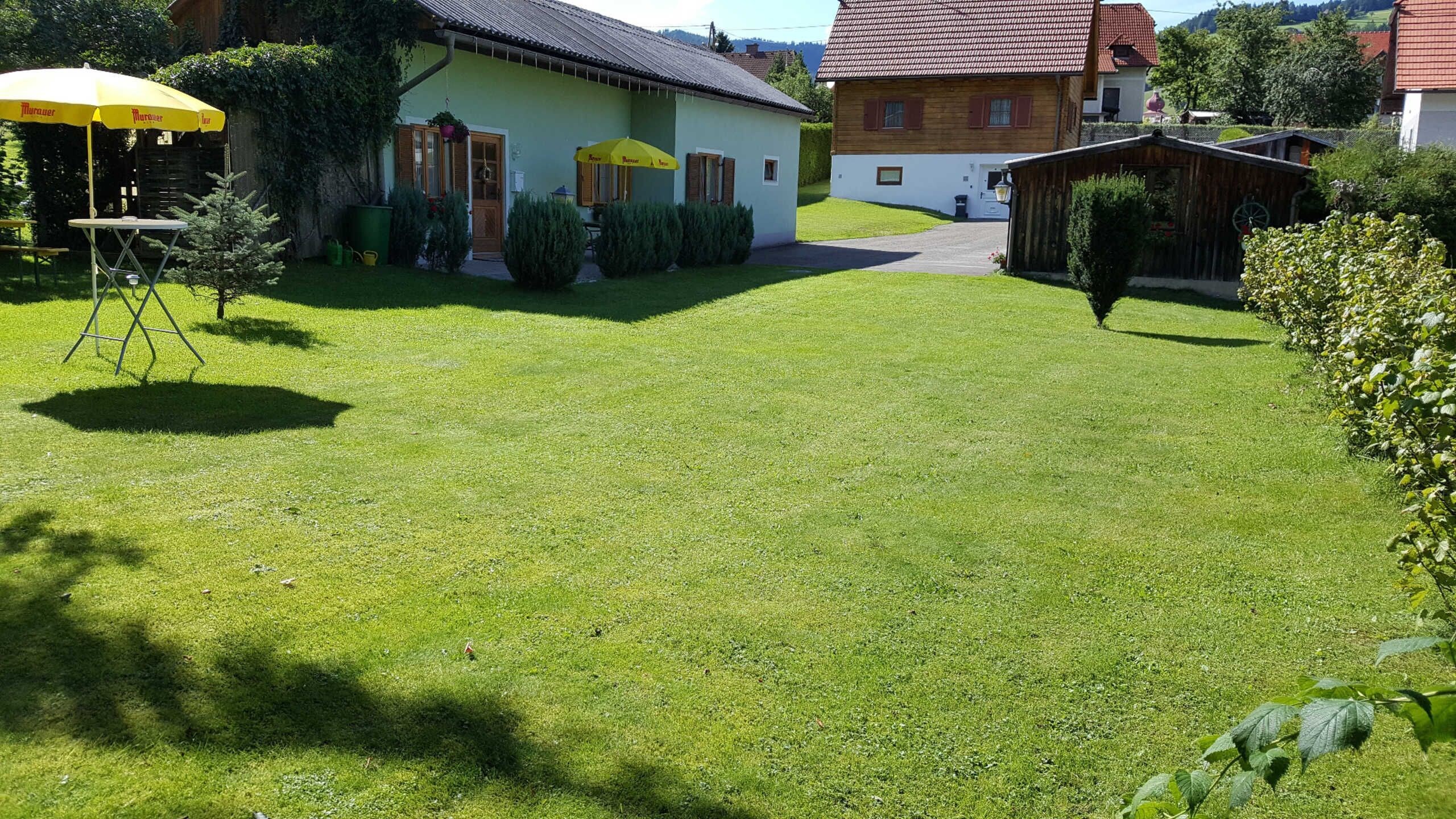 Une pelouse bien entretenue avec une petite table et des chaises sous un parasol jaune, une maison en bois avec un toit rouge et une clôture en bois.