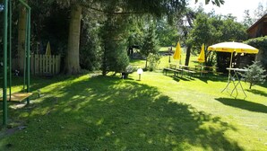 A grassy outdoor area with picnic tables and yellow umbrellas, surrounded by trees and a wooden fence. - Holiday home Findling Summer - House Findling (Sankt Lambrecht)