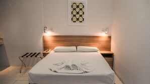 Una cama bien hecha con una presentación de toallas, cabecero de madera, mesitas de noche y una lámpara fijada a la pared.