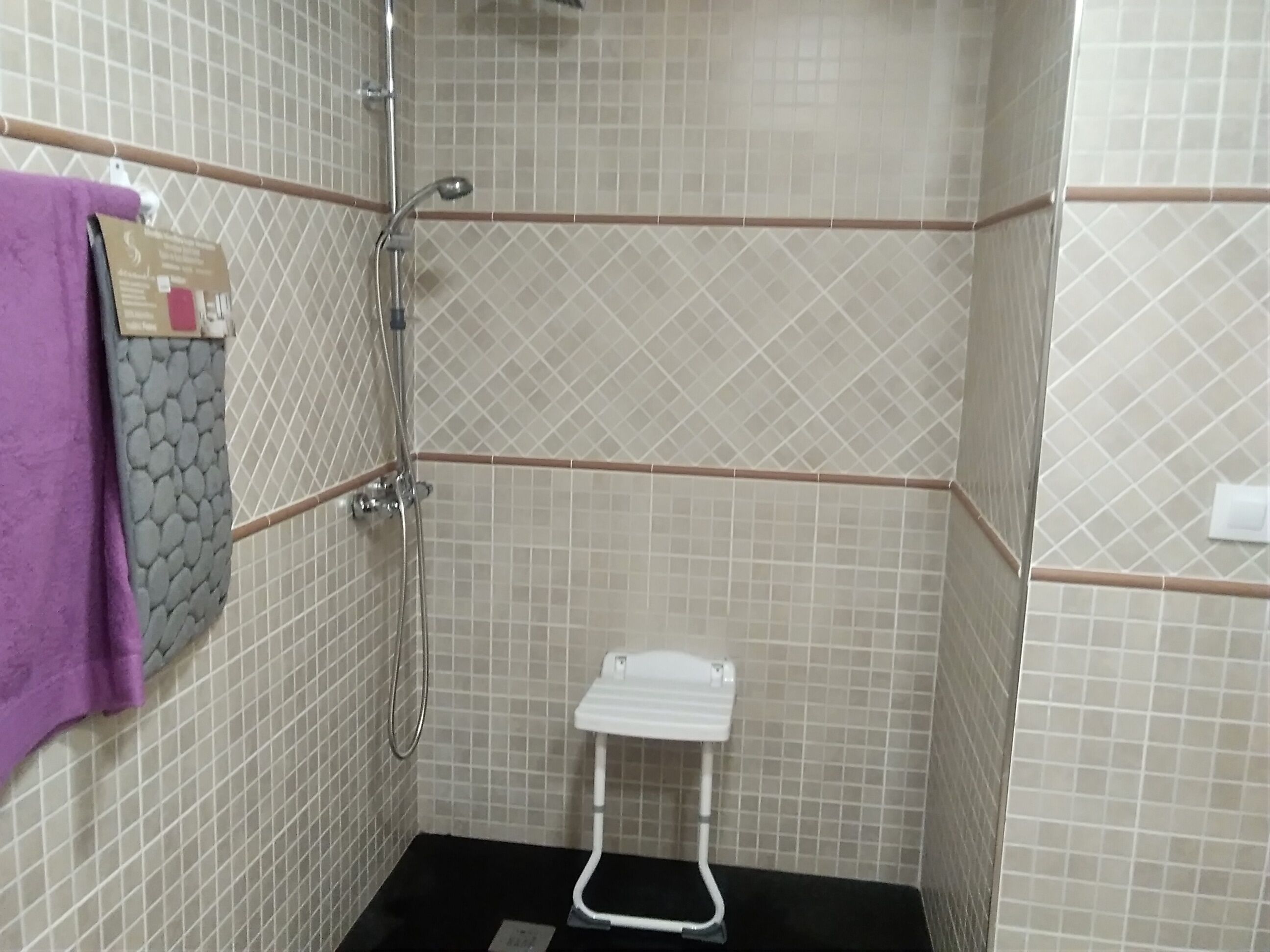 Un pequeño espacio de ducha con azulejos, un cabezal de ducha manual blanco, un taburete blanco y una toalla morada colgada en la pared.