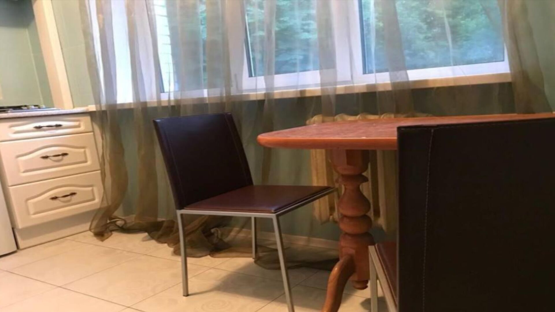 Ein Zimmer mit einem Holztisch, einem Stuhl, einem kleinen weißen Schrank und einem Fenster mit transparenten Vorhängen.