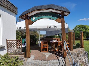 An outdoor seating area with wooden tables and benches, a covered entrance with a sign, and a white tent with 'VELTINS' branding. - !NEW! Holiday house Trudi, right on the Rothaarsteig, skiing, hiking or cycling (Winterberg)