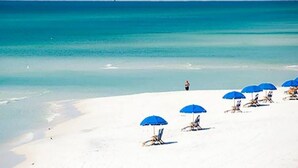 Een strand met wit zand en helderblauw water. Er staan diverse blauwe parasols en strandstoelen.