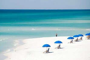 Ein Strand mit weißem Sand und klarem, blauem Wasser. Mehrere blaue Sonnenschirme und Liegen sind vorhanden.