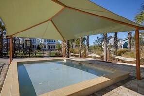 Una piscina con zona sombreada, rodeada de palmeras y casas.