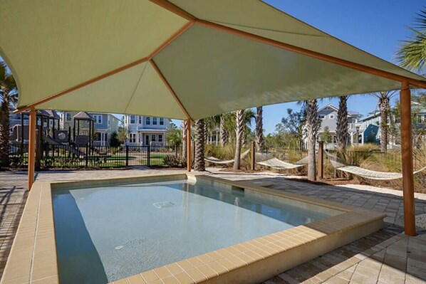 Una piscina con zona sombreada, rodeada de palmeras y casas.