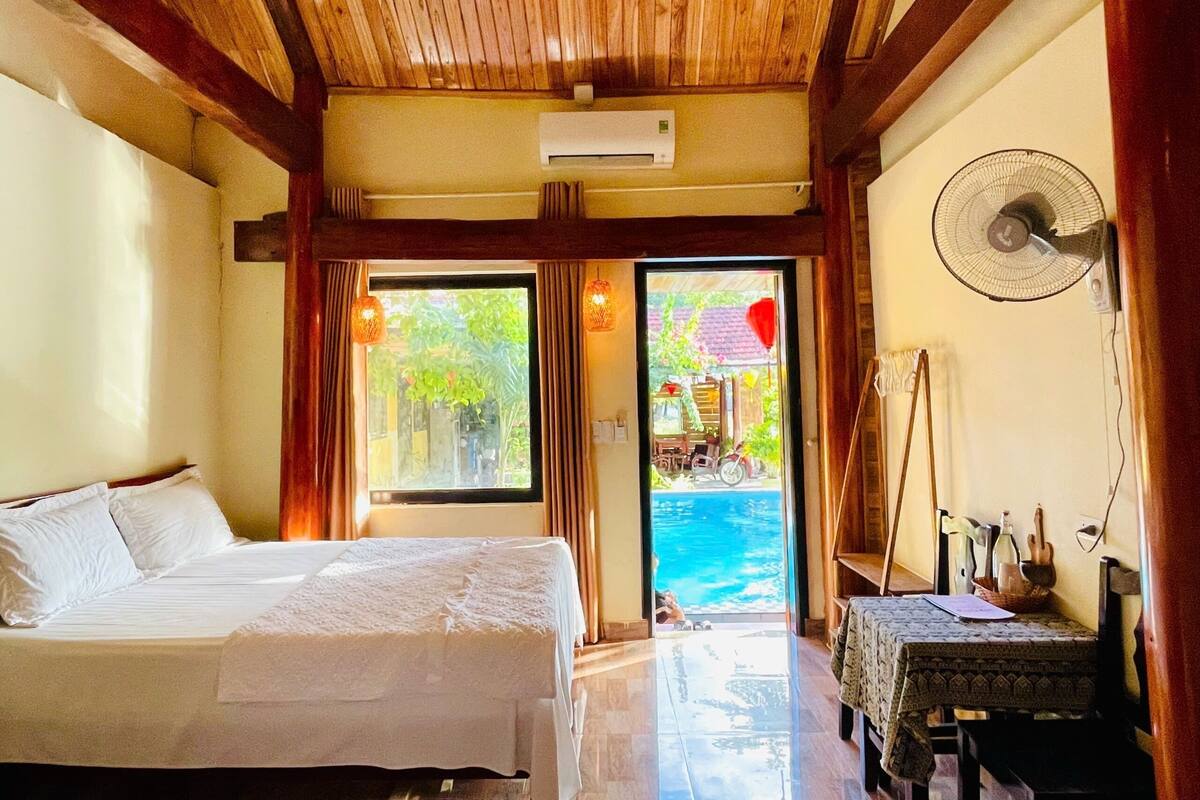 A hotel room with two beds, a fan, a table with a basket, and a view of a swimming pool and outdoor seating area.