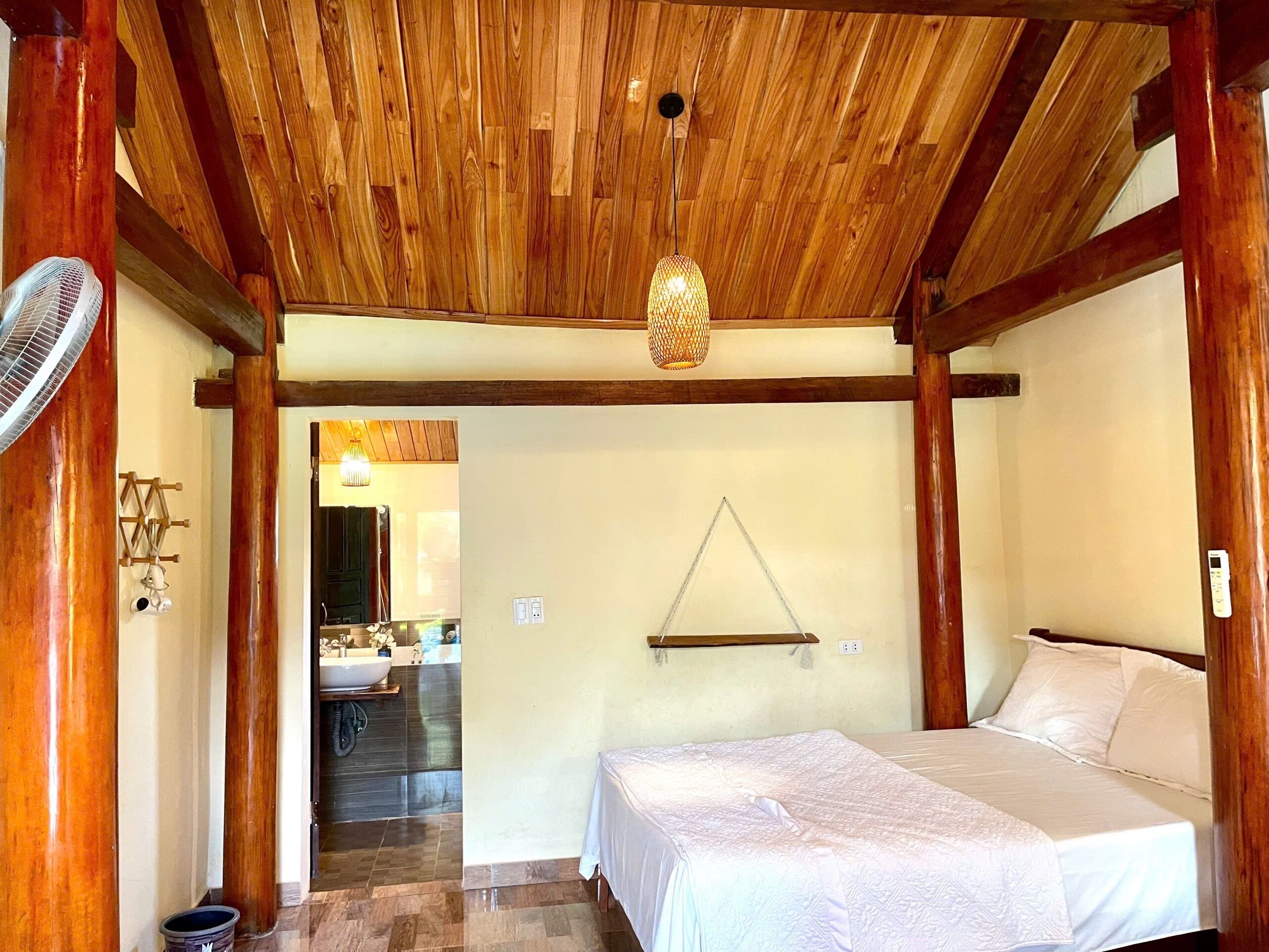 A bedroom with a wooden ceiling, a bed, a bedside table, a hanging light fixture, and a view into the bathroom.