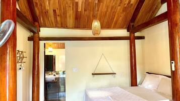 A bedroom with a wooden ceiling, a bed, a bedside table, a hanging light fixture, and a view into the bathroom.