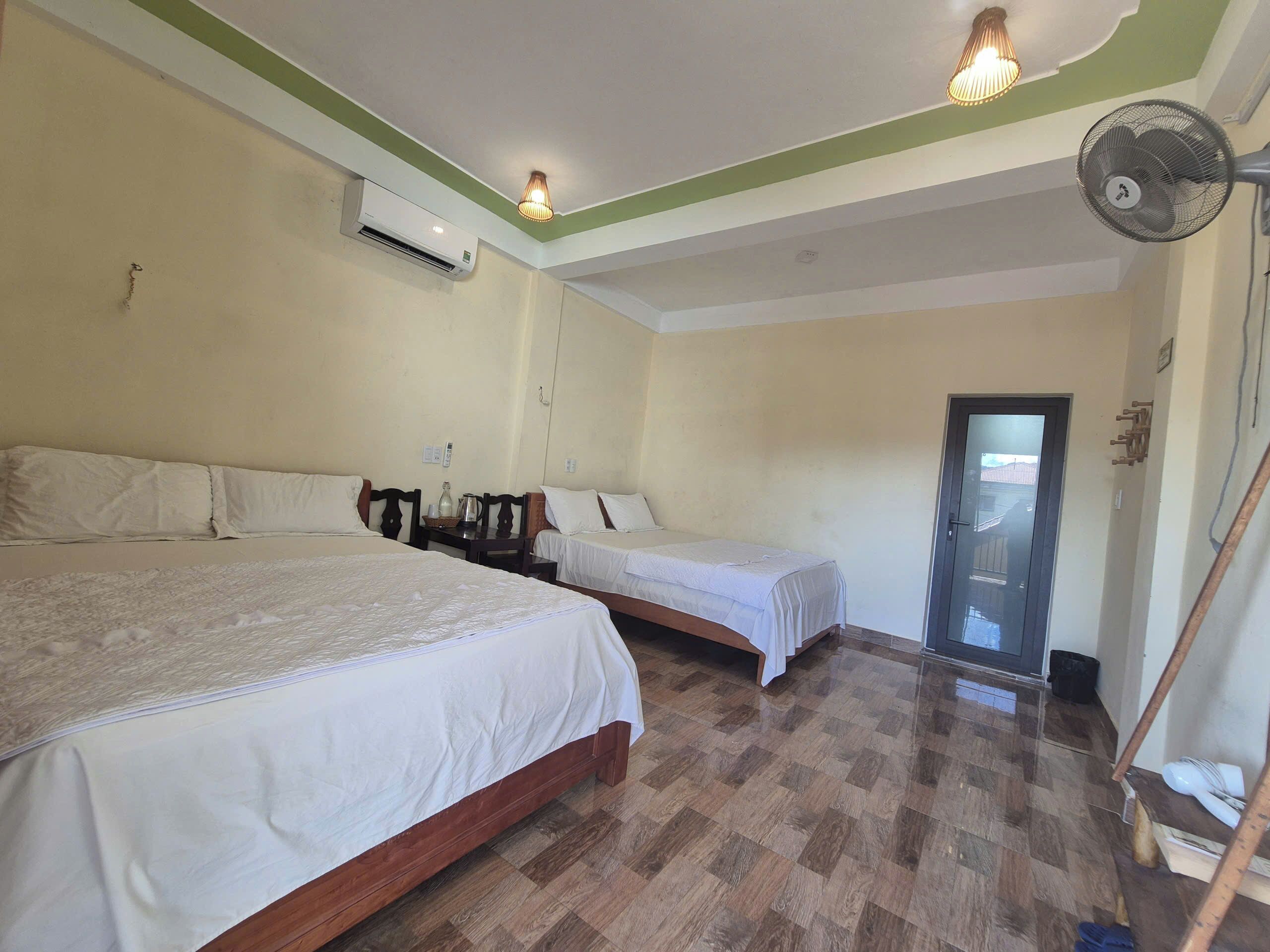 Habitación de hotel con dos camas individuales, piso de madera, unidad de aire acondicionado y ventilador de techo.