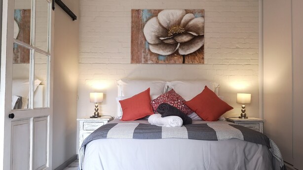 A bedroom with a large bed, two bedside tables with lamps, a white brick wall, and a large floral painting above the bed. - Self Catering Fully Equipped Cottage set in a leafy suburb (Plumstead)