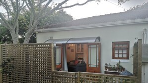 A house with a wooden fence, a door leading to a living area with a sofa, and a window with plants on the right. - Self Catering Fully Equipped Cottage set in a leafy suburb (Plumstead)