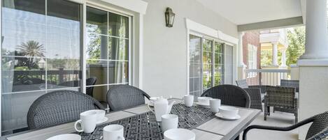 A dining area with a table set for four, surrounded by wicker chairs, large windows, and a view of a building in the background.