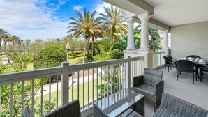 A covered outdoor patio with wicker furniture, white railings, and palm trees in the background.