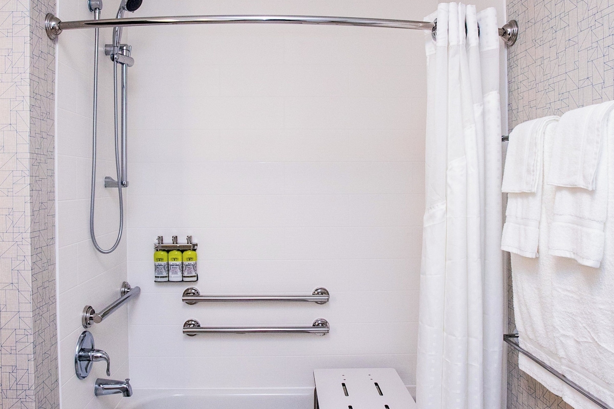 A shower with a handheld showerhead, a handheld shower attachment, a handheld shower hose, a grab bar, a towel rack, and a small step stool.