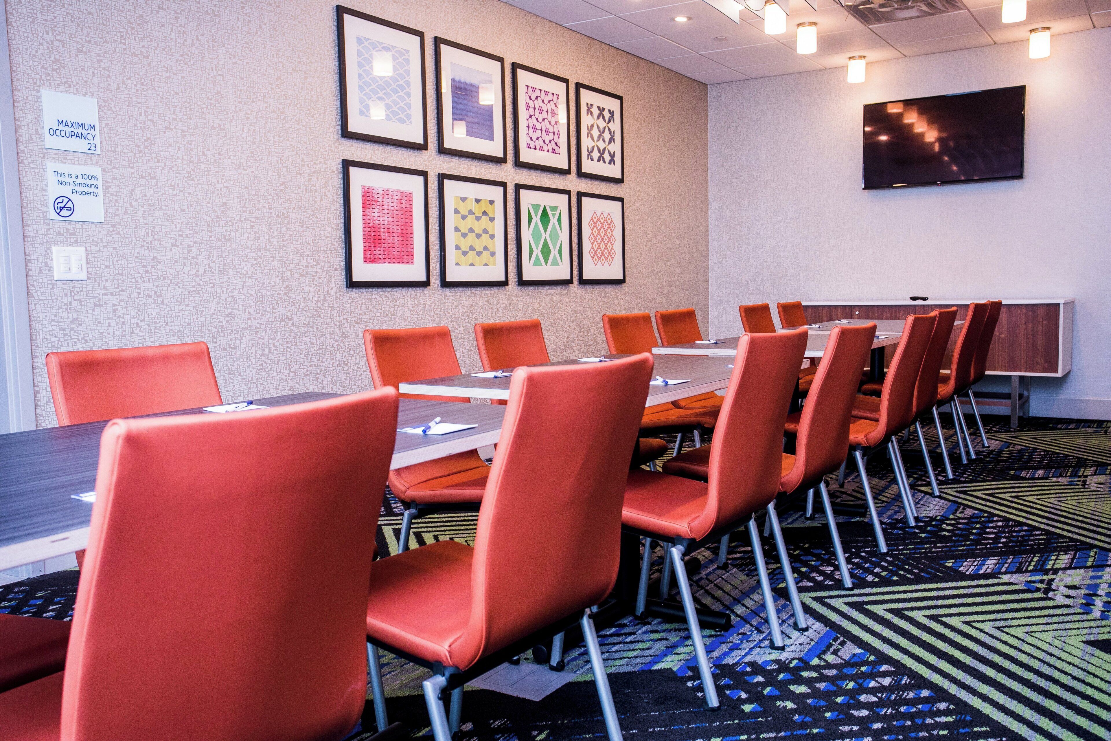 A conference room with a long table and orange chairs, framed artwork on the wall, and a flat-screen TV.
