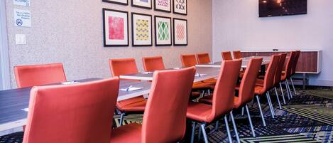 A conference room with a long table and orange chairs, framed artwork on the wall, and a flat-screen TV.