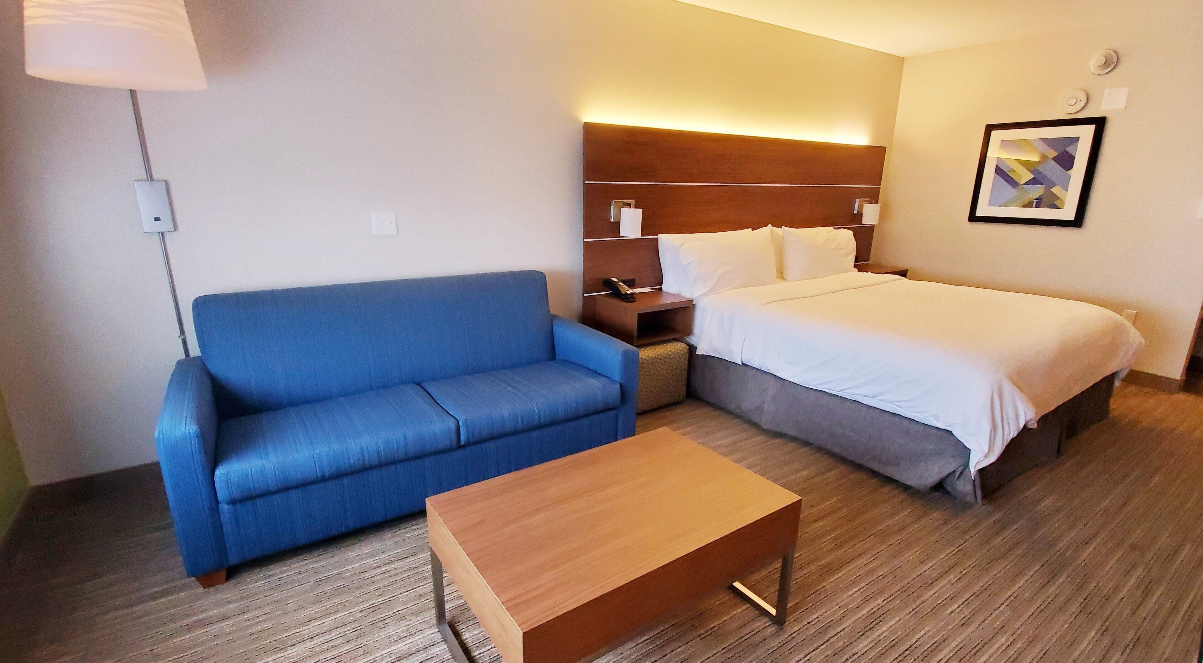 A hotel room with a blue sofa, a wooden coffee table, a bed with white bedding, a nightstand, and a wall-mounted lamp.