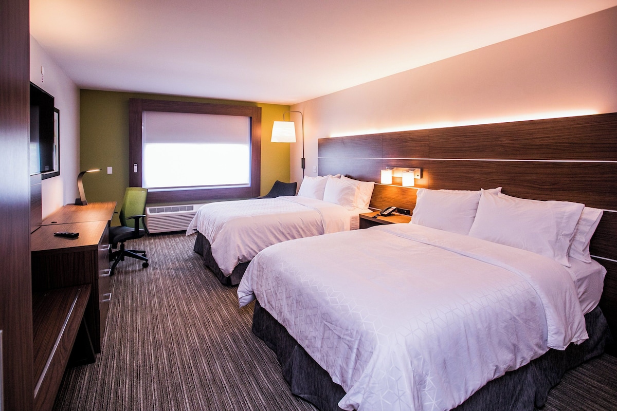 A hotel room with two beds, a desk, a chair, a TV, and a window with blinds.