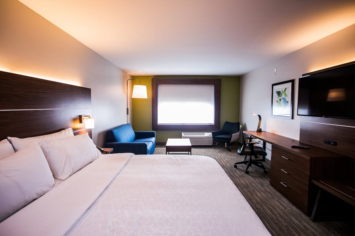 A hotel room with a large bed, a desk, a chair, a TV, and a window with blinds.
