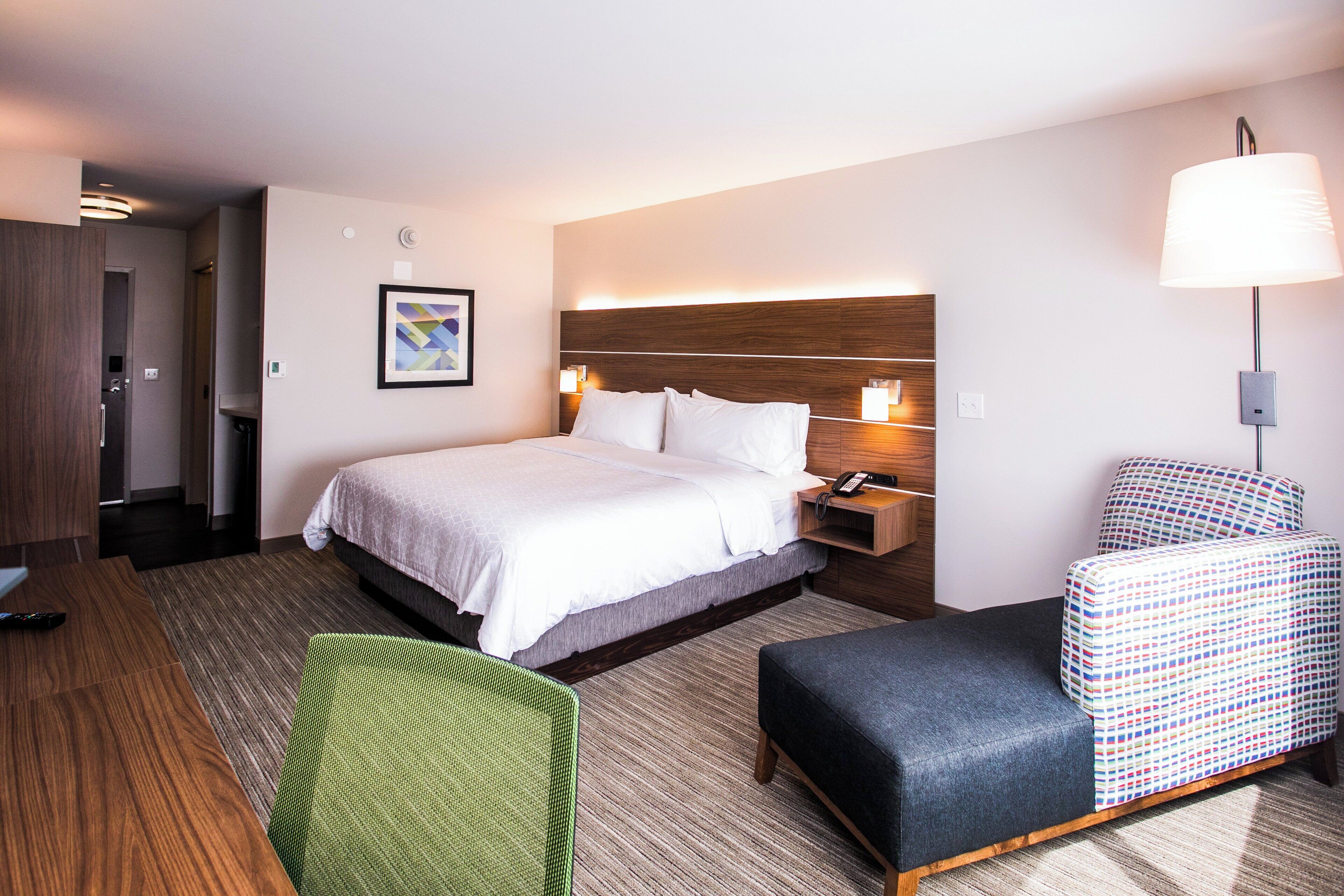 Una habitación de hotel con cama, mesita de noche, lámpara, silla y un cuadro en la pared.